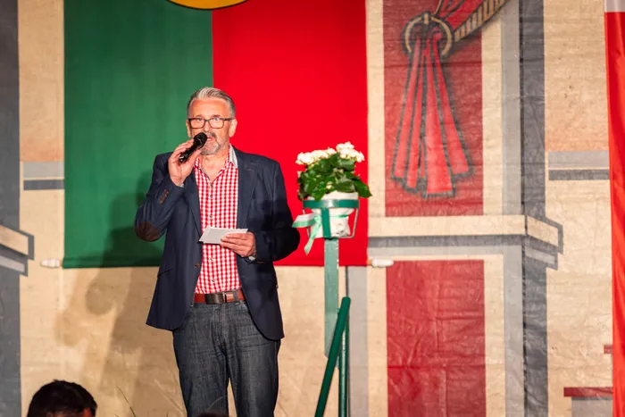 Ein Mann im blauen Sakko und rot-weiß kariertem Hemd spricht in ein Mikrofon vor großen roten und grünen Bannern.