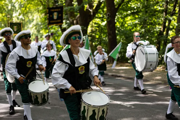 Eine Spielmannszug marschiert auf einer Straße, die Trommler tragen grüne und weiße Uniformen und spielen auf ihren Trommeln.