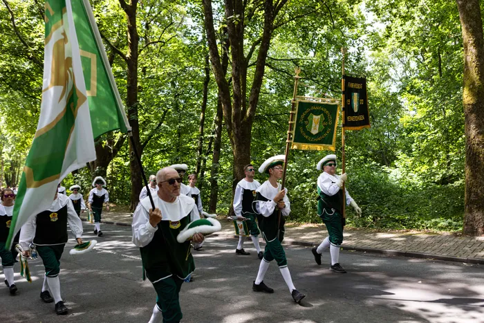 Eine Schützengilde marschiert auf einer Straße, vorneweg ein Mann mit einer Flagge und weitere Mitglieder mit einem Banner.