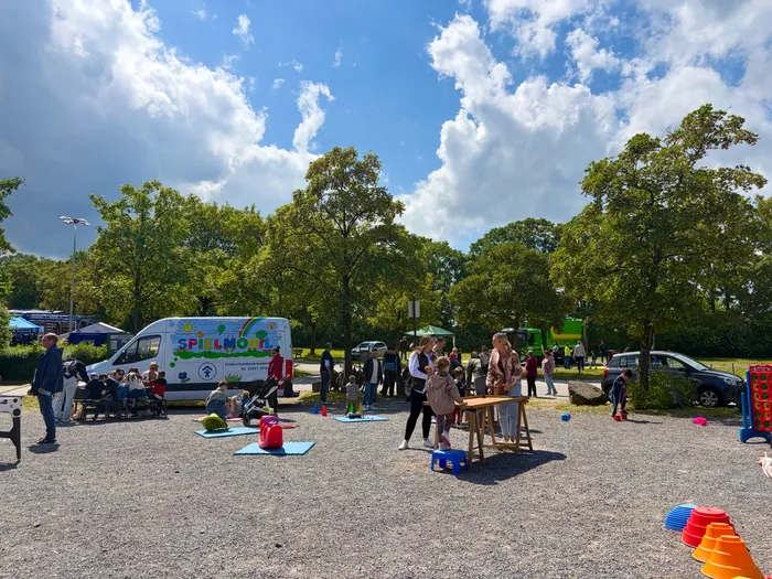 Menschen jeden Alters sind auf einem Parkplatz versammelt, wo ein bunt bemalter Spielmobil-Transporter steht. Im Hintergrund sind Bäume und geparkte Autos zu sehen, und Kinder spielen auf blauen Matten.
