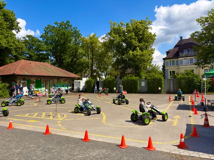 Kinder und Jugendliche fahren auf einem abgesperrten Platz mit kleinen Elektro-Karts. Im Hintergrund ein Gebäude mit rotem Dach und ein Wohnhaus. Verkehrskegel markieren eine Rennstrecke.