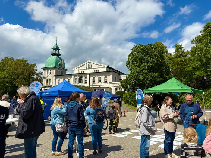 Menschen stehen vor Zelten mit dem Logo der Polizei und der Verkehrswacht, im Hintergrund ein helles Gebäude mit Kuppel und Bäume unter blauem Himmel. Der Aktionstag der Verkehrswacht Lüdenscheid lockt viele Besucher an.