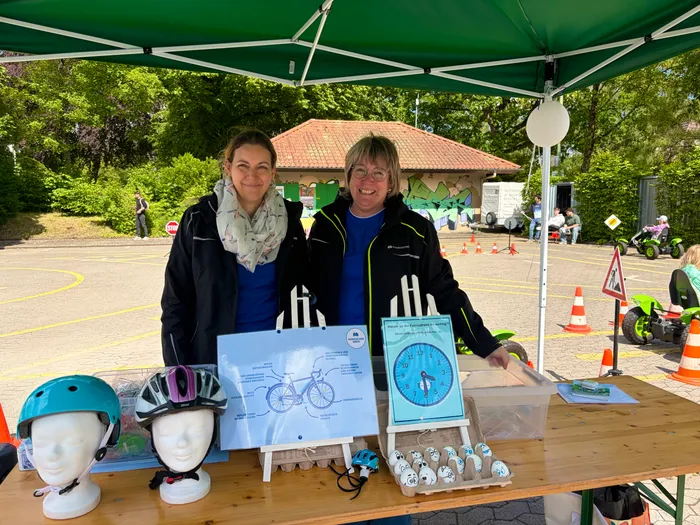 Zwei Frauen stehen an einem Infostand mit Fahrradhelmen und Informationsmaterialien zum Thema Verkehrssicherheit. Im Hintergrund sind weitere Menschen und ein grüner Pavillon zu sehen.