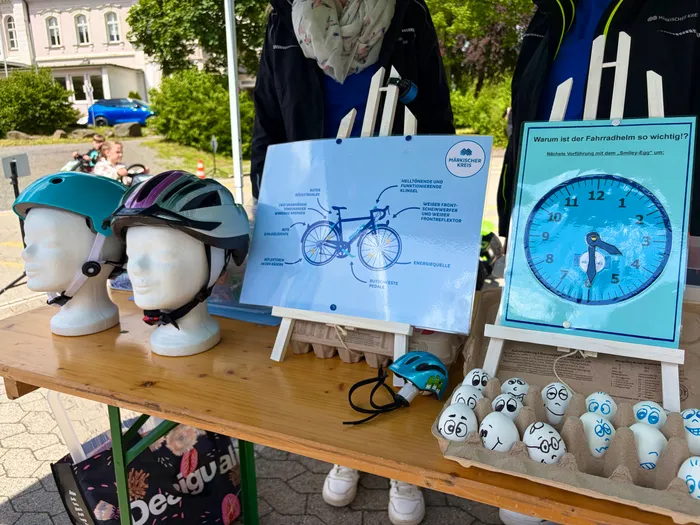 Ein Tisch zeigt Fahrradhelme, Informationsmaterialien zur Fahrradsicherheit und Plüschfiguren. Im Hintergrund ist ein Plakat mit der Aufschrift „Warum ist der Fahrradhelm so wichtig?“ zu sehen.