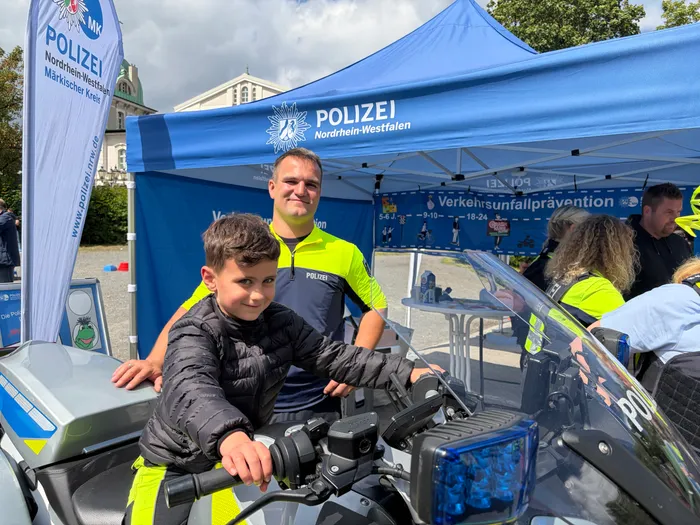 Ein Junge sitzt auf einer Polizeimotorrad und lächelt in die Kamera. Im Hintergrund steht ein Zelt mit dem Aufdruck „Polizei Nordrhein-Westfalen“ und Mitarbeiter der Verkehrswacht.
