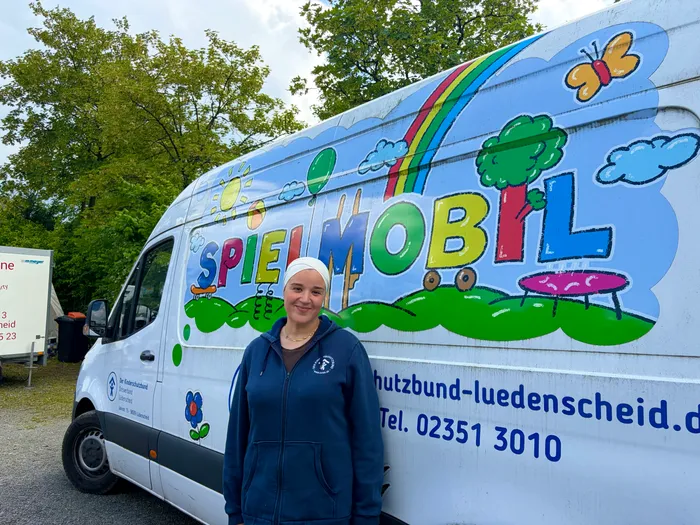 A woman stands in front of a white van decorated with a rainbow, butterflies, and the word 'SPIELMOBIL'. The van displays contact information for the 'Verkehrswacht Lüdenscheid'.