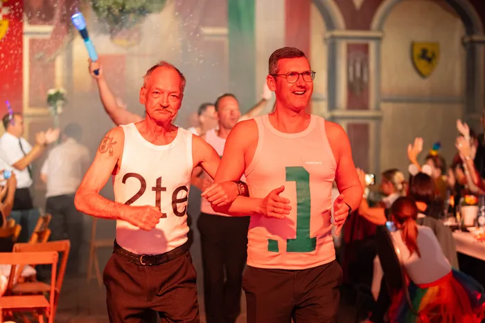 Zwei Männer in weißen Tanktops mit den Nummern „2te“ und „1“ tanzen bei einem Schützenfest.