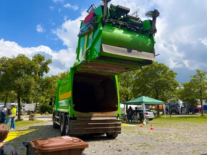 Ein grüner Müllwagen mit geöffnetem Ladebereich steht auf einem Platz, umgeben von Besuchern und einem Infostand. Im Vordergrund ist ein brauner Müllcontainer zu sehen.
