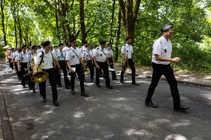 Eine Musikgruppe schreitet auf einem Weg durch einen Wald. Vorneweg geht ein Mann mit einer Tuba, gefolgt von weiteren Musikern mit verschiedenen Blasinstrumenten.