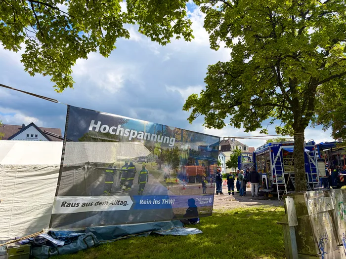 Großes Banner mit dem Schriftzug 'Hochspannung' und Bildern von Einsatzkräften der Feuerwehr und des THW vor einer Wohnbebauung und Einsatzfahrzeugen. Darunter steht 'Raus aus dem Alltag. Rein ins THW!'.