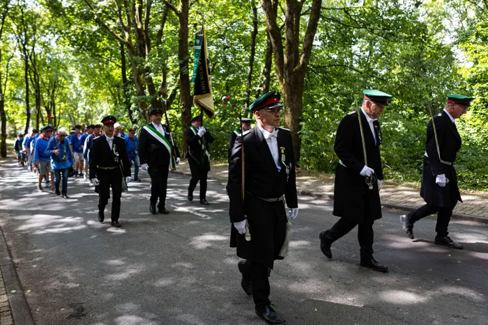 Schützen marschieren in dunklen Uniformen und mit grünen Mützen auf einem Weg unter Bäumen, eine Fahne ist zu sehen.