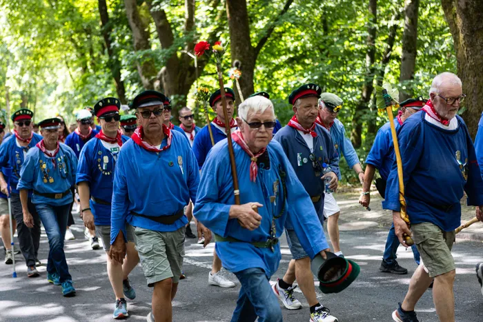 Schützen ziehen bei einem Festumzug durch eine von Bäumen gesäumte Straße. Viele tragen eine blaue Uniform mit rotem Tuch und eine grüne Schützenmütze.