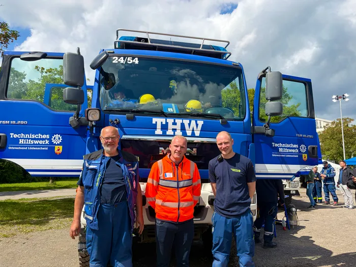 Drei Männer in Uniformen stehen vor einem blauen THW-Fahrzeug mit der Aufschrift 