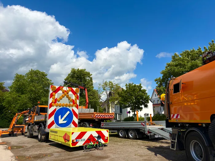 Ein orangefarbener Abschleppwagen mit einem Anhänger steht auf einer Straße. Auf dem Anhänger ist ein blaues Schild mit einem Pfeil nach links und ein Kinderfahrrad zu sehen. Im Hintergrund sind Bäume und Häuser zu erkennen.