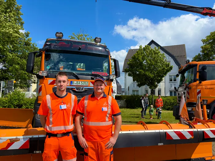 Zwei Männer in orangefarbenen Arbeitskleidern stehen vor einem großen orangefarbenen Abschleppwagen mit der Aufschrift 