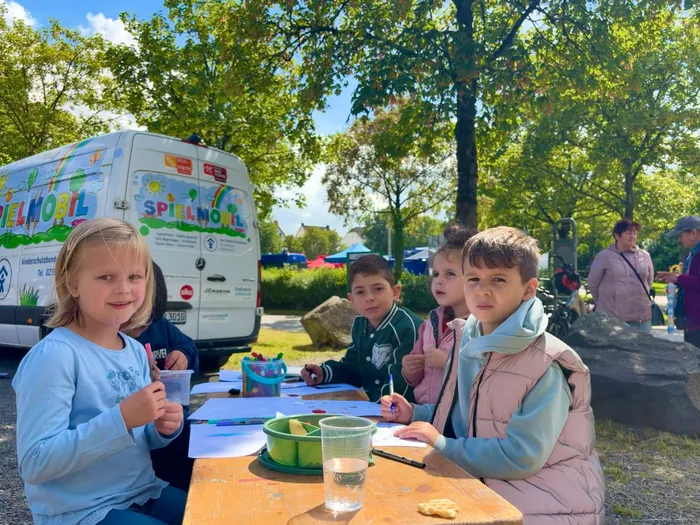 Kinder sitzen an einem Tisch und malen, während im Hintergrund ein bunt bemalter Spielmobil-Bus steht.