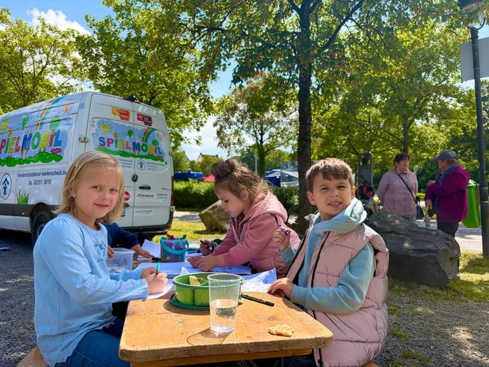 Drei Kinder sitzen an einem Holztisch und malen, während im Hintergrund ein bunt bemalter Spielmobil-Transporter steht. Der Aktionstag der Verkehrswacht Lüdenscheid bietet offenbar kreative Beschäftigung für Kinder.