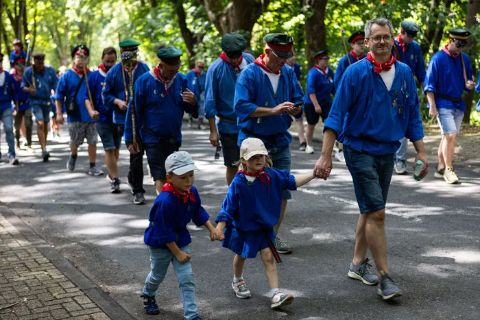 Schützen ziehen bei einem Festumzug durch eine parkähnliche Landschaft. Im Vordergrund halten ein Mann und ein Kind Hände, beide tragen Schützenuniformen.