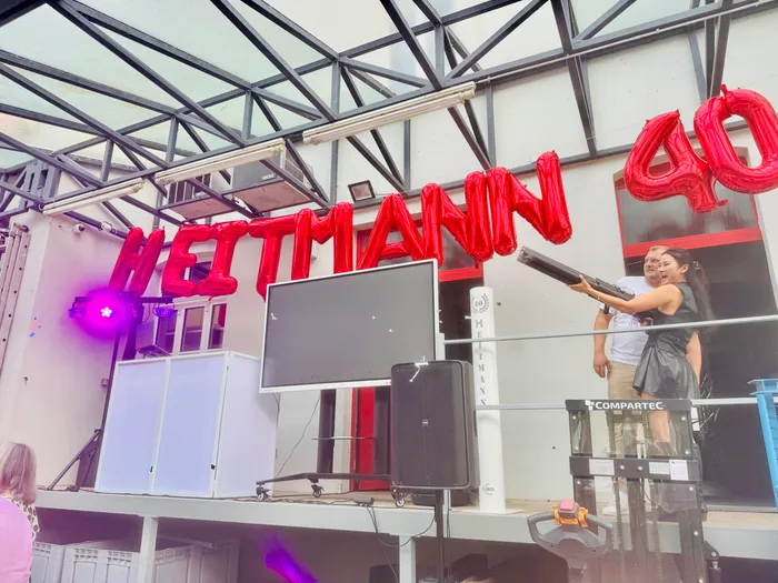 Rote Folienballons mit der Aufschrift „HEITMANN 40“ unter einem Glasdach auf einer Terrasse mit technischem Equipment und zwei Personen.