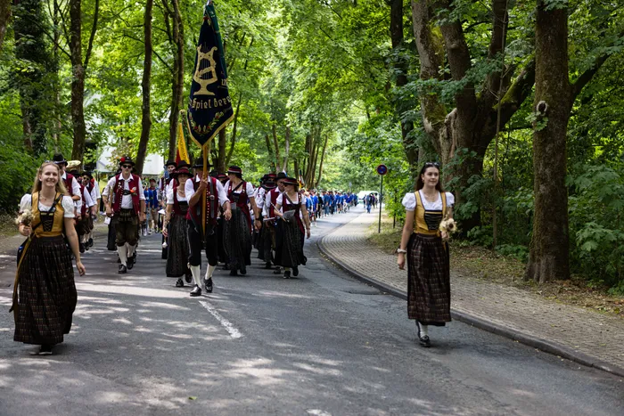 Eine Gruppe von Menschen in traditioneller Tracht marschiert auf einer Straße, eine Person trägt eine Fahne mit dem Schriftzug „Spielberg“.