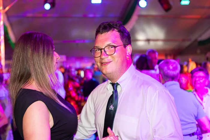 Ein lächelnder Mann mit Brille und weißem Hemd steht bei einer festlichen Veranstaltung mit bunter Beleuchtung.