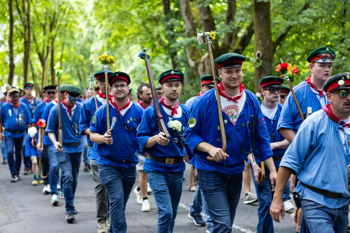 Schützen ziehen bei einem Festumzug durch eine von Bäumen gesäumte Straße. Viele tragen blaue Jacken, Jeans und Mützen.