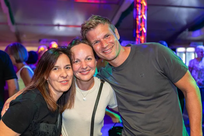 Zwei Frauen und ein Mann lächeln eng beieinander posierend vor einem Hintergrund mit bunten Lichtern beim Schützenfest.