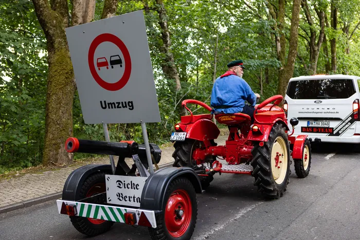 Ein roter Oldtimer-Traktor fährt auf einer Straße, beladen mit einem Schild mit einem Umzugsverbot und einem weiteren Schild mit der Aufschrift „Dicke Berta“. Ein Mann mit Kappe sitzt auf dem Traktor. Im Hintergrund ein weißer Van und grüße Bäume.