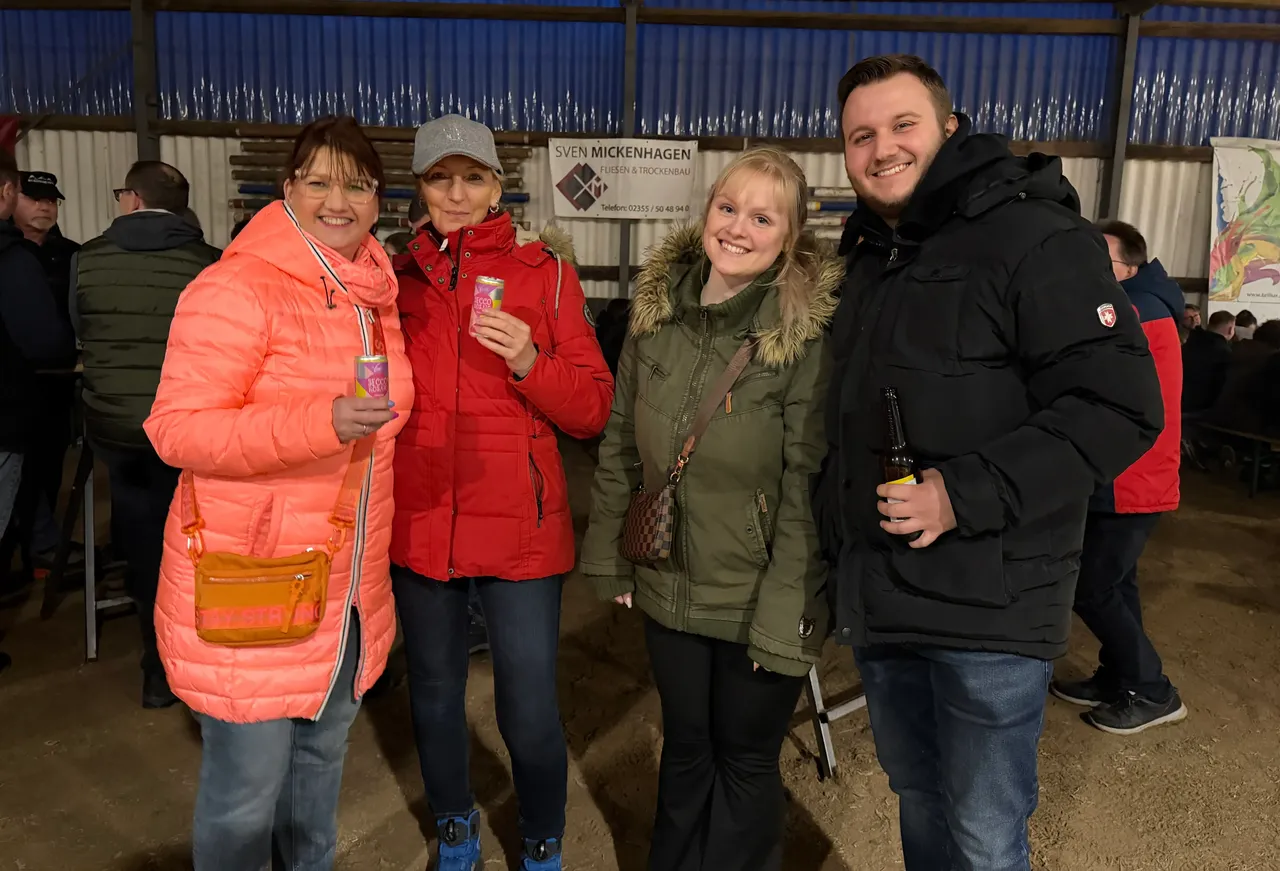 Vier lächelnde Frauen und ein Mann stehen eng zusammen und halten Getränke in der Hand. Im Hintergrund ein Banner mit dem Namen „Sven Mickenhagen“ und weitere Besucher eines Osterfeuers.