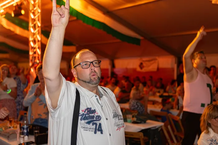 Ein Mann mit Brille hebt im Festzelt beim Schützenfest die Hand, während im Hintergrund Gäste an Tischen sitzen.