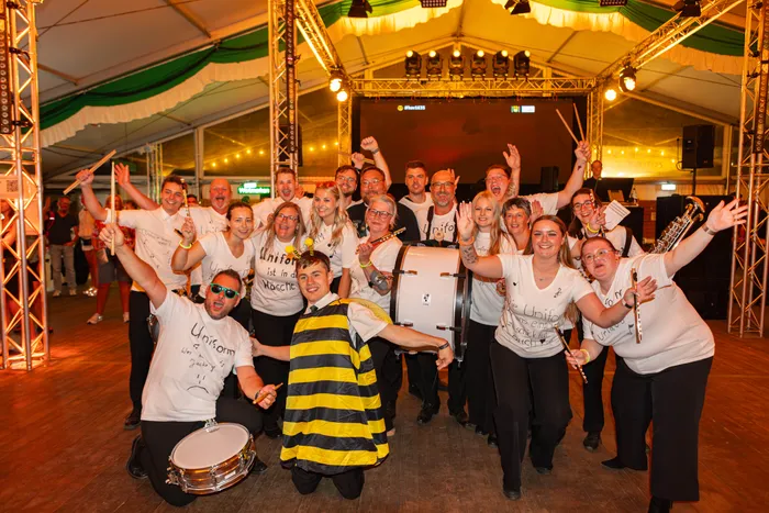 Eine Gruppe von Menschen in weißen T-Shirts mit Musikinstrumenten posiert fröhlich unter einem Festzelt, eine Person trägt ein Bienenkostüm.
