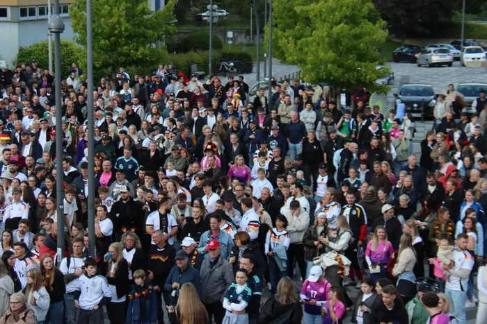 Eine große Menschenmenge jubelt auf dem Otto-Fuchs-Platz, viele tragen Deutschlandtrikots und schwenken Flaggen. Die Stimmung ist ausgelassen, da die deutsche Nationalmannschaft gegen Curaçao gewonnen hat.