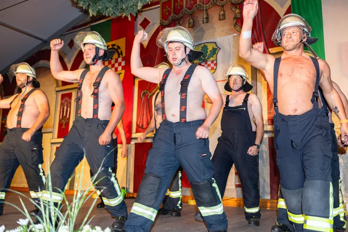 Mehrere Männer in Feuerwehrhosen und Helmen tanzen oberkörperfrei auf einer Bühne.