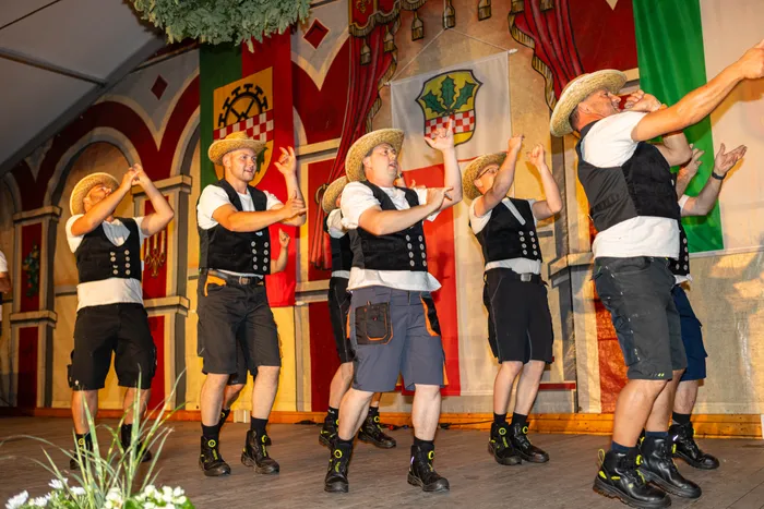 Männer in traditioneller Tracht mit Lederhosen und Hüten tanzen auf einer Bühne vor bunten Fahnen.