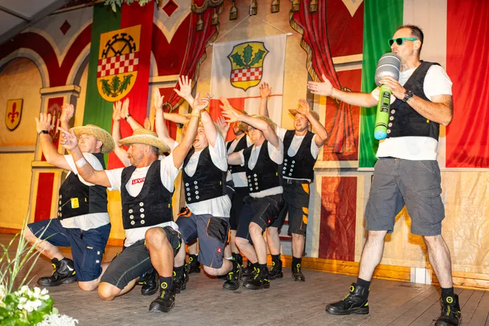 Männer in Trachten mit Strohhüten tanzen auf einer Bühne vor bunten Bannern während eines Schützenfests.