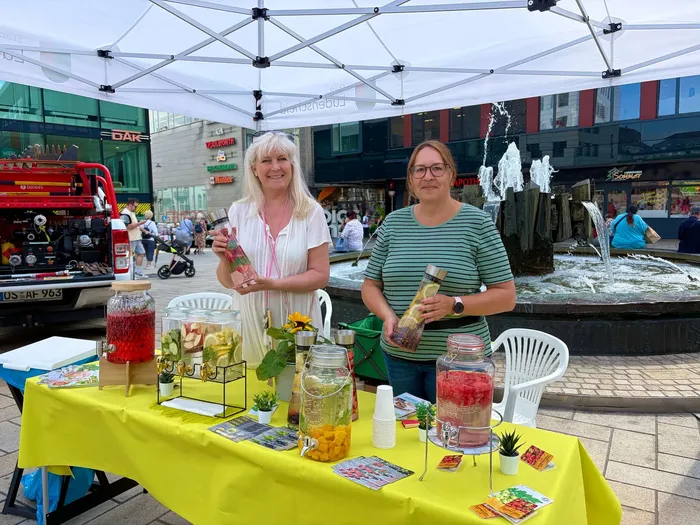 Anja Weber und Susanne Löffler stehen an einem Stand mit verschiedenen Glaskaraffen voller Fruchtwasser unter einem weißen Sonnendach.