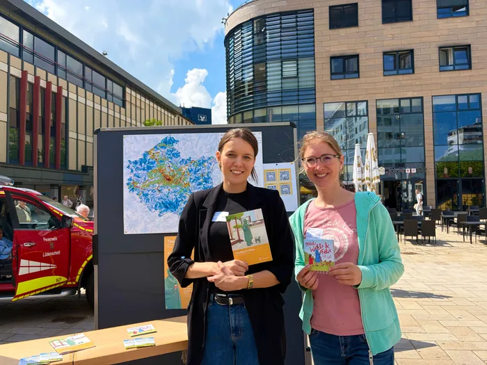 Zwei Frauen stehen vor einer Infostelle mit Plakaten und Broschüren zum Thema Hitzeschutz. Die Frau links hält ein Handbuch, die rechte ein Kinderbuch.