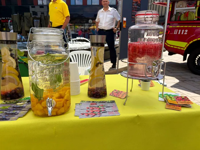 The image shows a table covered with a yellow cloth displaying several glass dispensers filled with colorful infused water. Flyers advertising the flavors are placed in front of the dispensers. In the background, people and a red vehicle are visible.