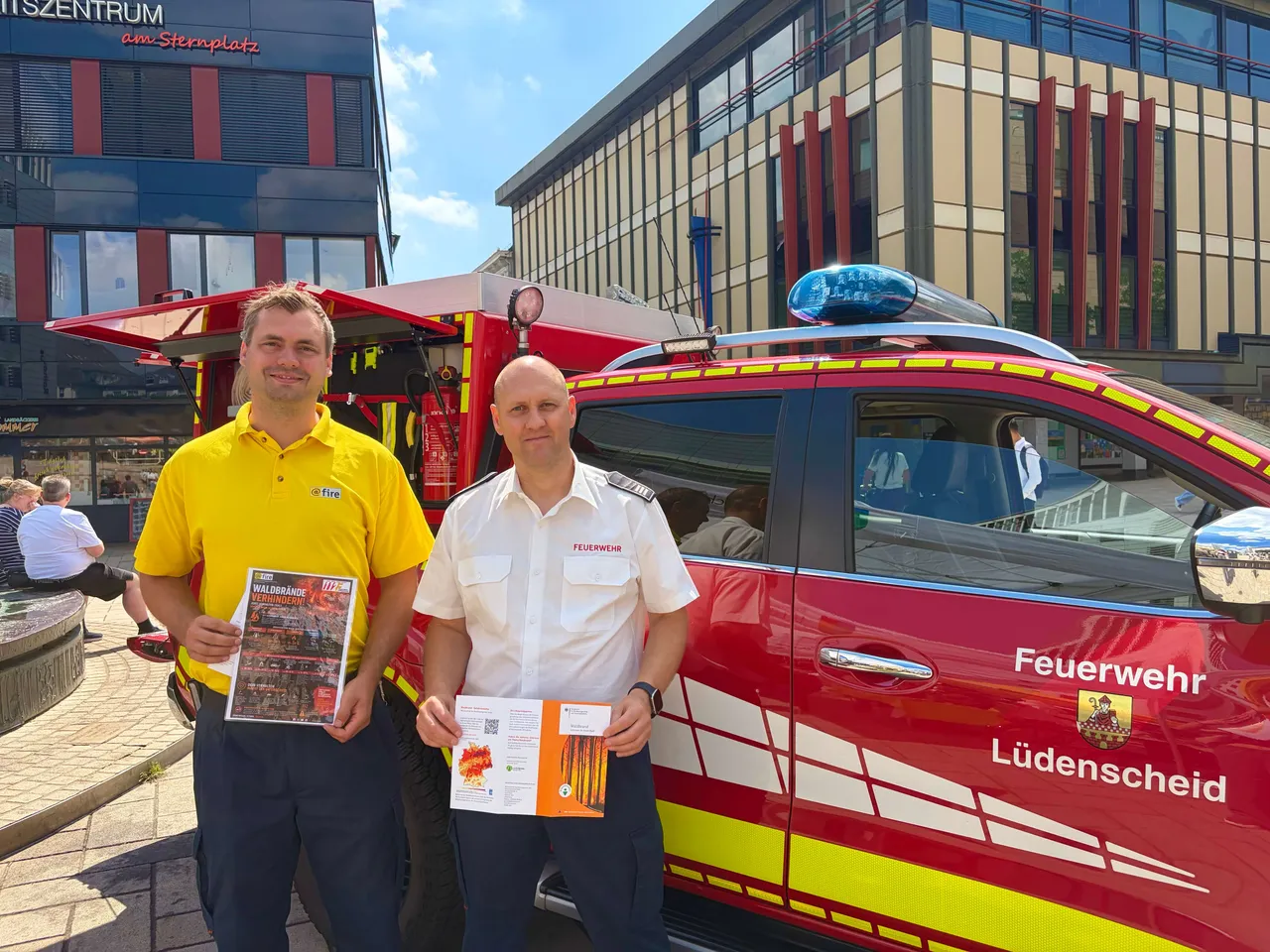 Zwei Feuerwehrleute in Uniformen halten Informationsmaterialien vor einem roten Feuerwehrauto und einem modernen Gebäude.