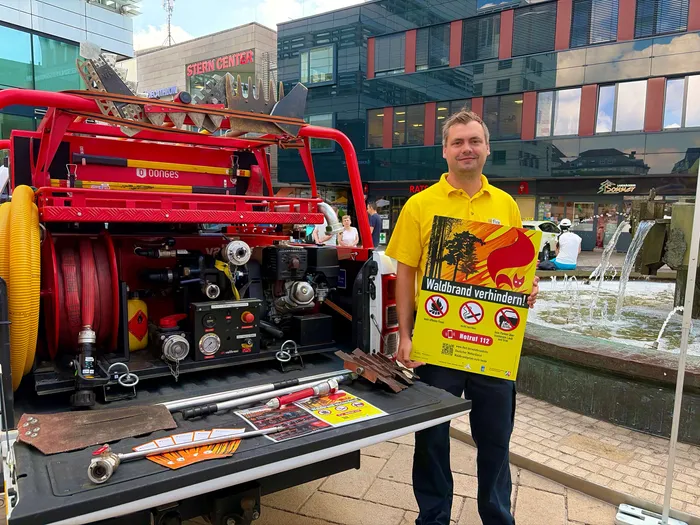 Sean Micke vom internationalen Katastrophenschutz steht vor einem roten Feuerwehrauto und hält ein Plakat mit der Aufschrift „Waldbrand verhindern!“ in die Kamera.