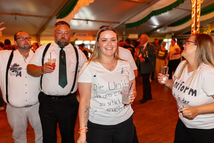 Eine lächelnde Frau in einem weißen T-Shirt mit Aufschrift hält ein Getränk, während Menschen bei einem Schützenfest in einem Festzelt feiern.