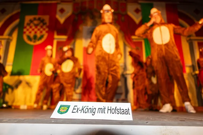 Personen in braunen Kostümen auf einer Bühne vor bunten Vorhängen; ein Schild im Vordergrund trägt die Aufschrift „Ex-König mit Hofstaat“.