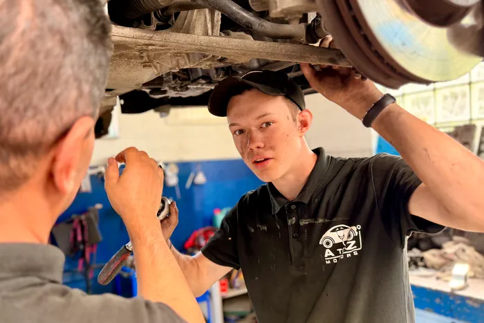 Julian Artmeier arbeitet gemeinsam mit einem Mechaniker an der Unterseite eines Fahrzeugs in einer Werkstatt.