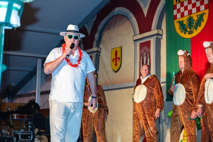 Ein Mann mit weißem Hut und rotem Blumenkranz singt in ein Mikrofon, während Personen in Bärenkostümen auf einer Bühne stehen.