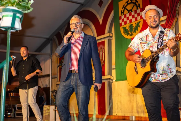 Ein Mann mit Brille und blauem Sakko spricht in ein Mikrofon auf einer Bühne beim Schützenfest, während ein Musiker mit Gitarre daneben steht.
