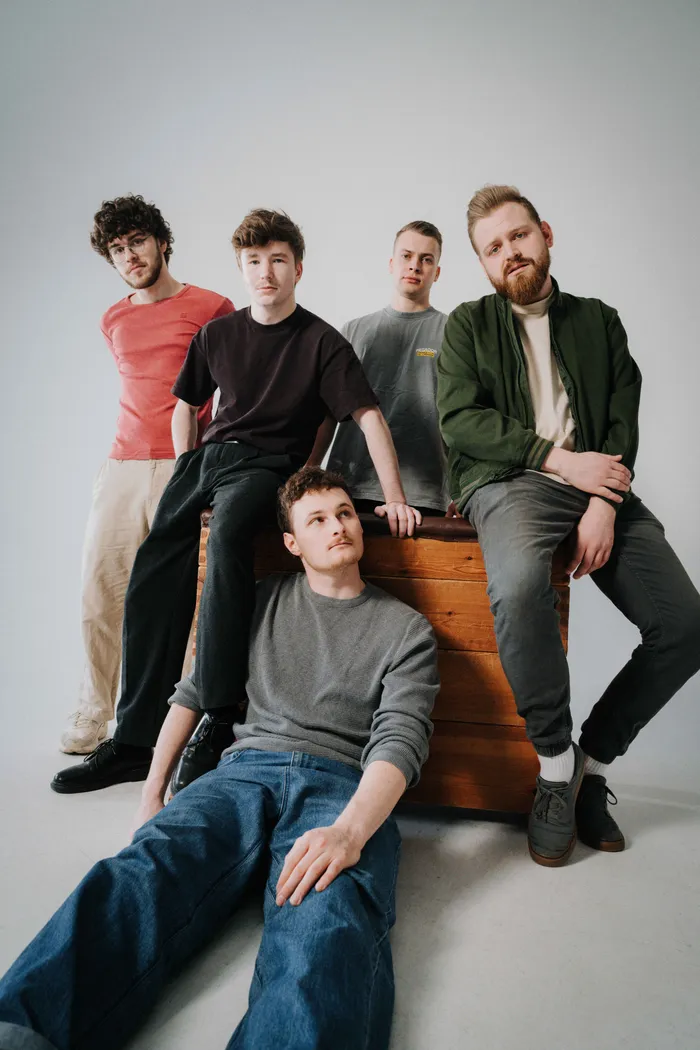 Fünf junge Männer posieren für ein Bandfoto. Sie sitzen und lehnen auf einer hölzernen Kiste, ein Mann liegt davor. Die Band 5Raumfenster spielt gitarrenlastigen Rock mit deutschen Texten.
