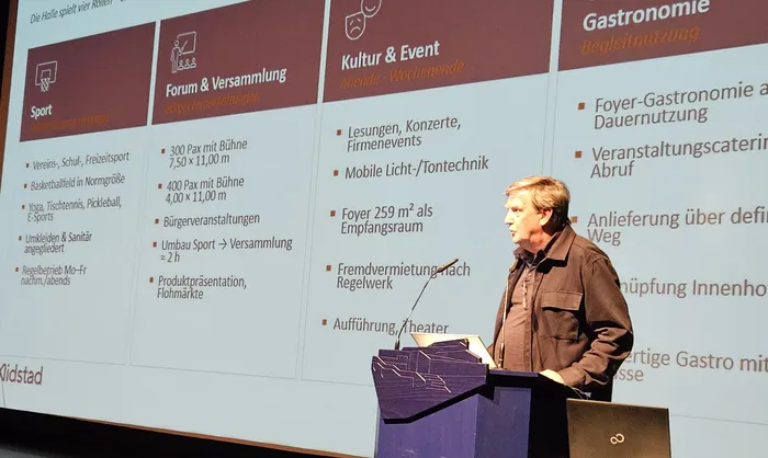 Architekt Christoph Irmisch steht an einem Rednerpult vor einer Projektionswand, die verschiedene Nutzungsbereiche eines geplanten Gebäudes zeigt.