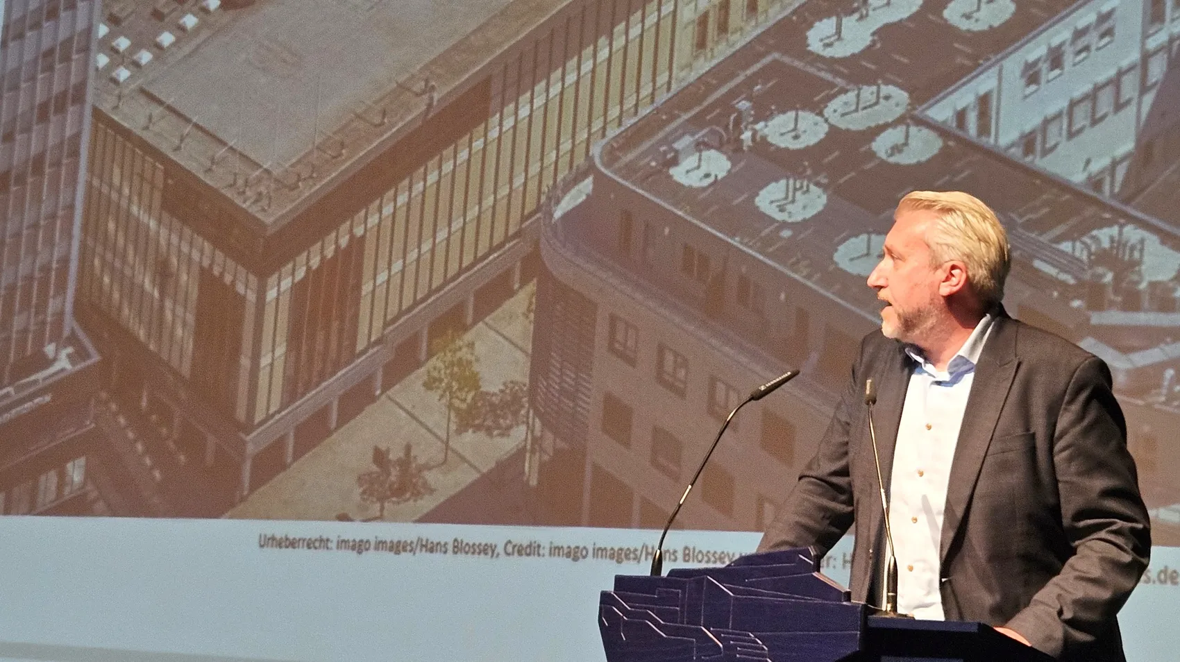Bürgermeister Sebastian Wagemeyer spricht an einem Rednerpult vor einer Projektion einer Luftaufnahme des P&C-Komplexes.