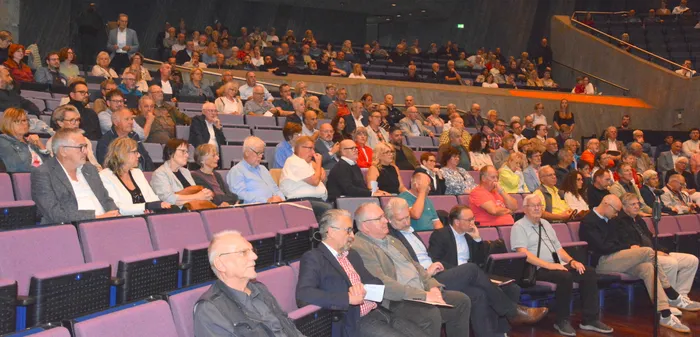 Zahlreiches Publikum sitzt in Reihen von lila Sitzen in einem großen Auditorium während einer Sitzung.