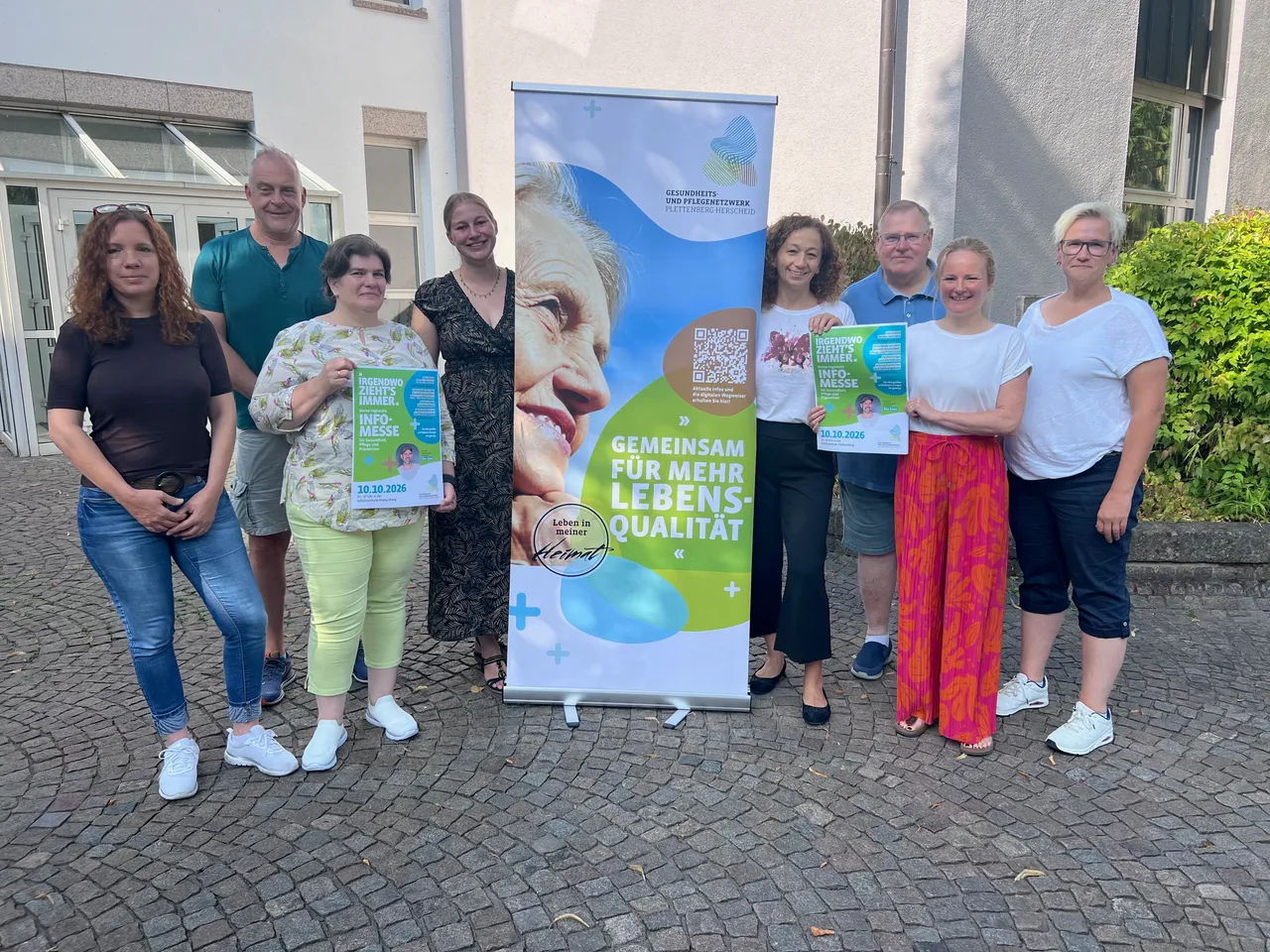 Eine Gruppe von Menschen steht um ein Werbebanner für die Infomesse „Irgendwo zieht's immer“ und hält Flyer in den Händen.